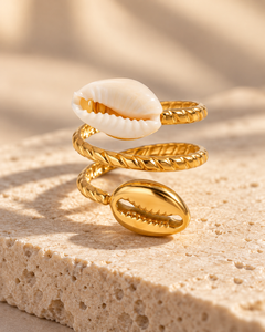 Bague COURA à spirale dorée torsadée, cauri blanc et cauri doré, posée sur du travertin beige.