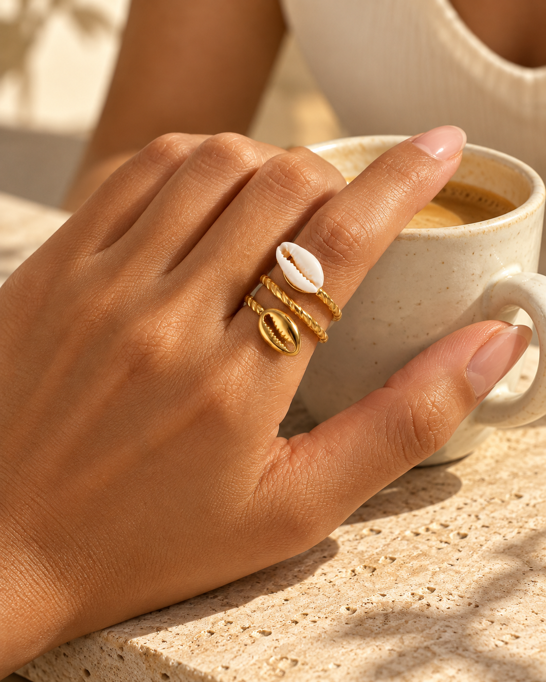 Bague COURA à deux cauris portée à l’index près d’une tasse de café, en lumière naturelle.