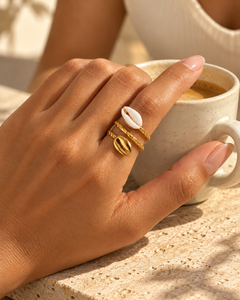 Bague COURA à deux cauris portée à l’index près d’une tasse de café, en lumière naturelle.