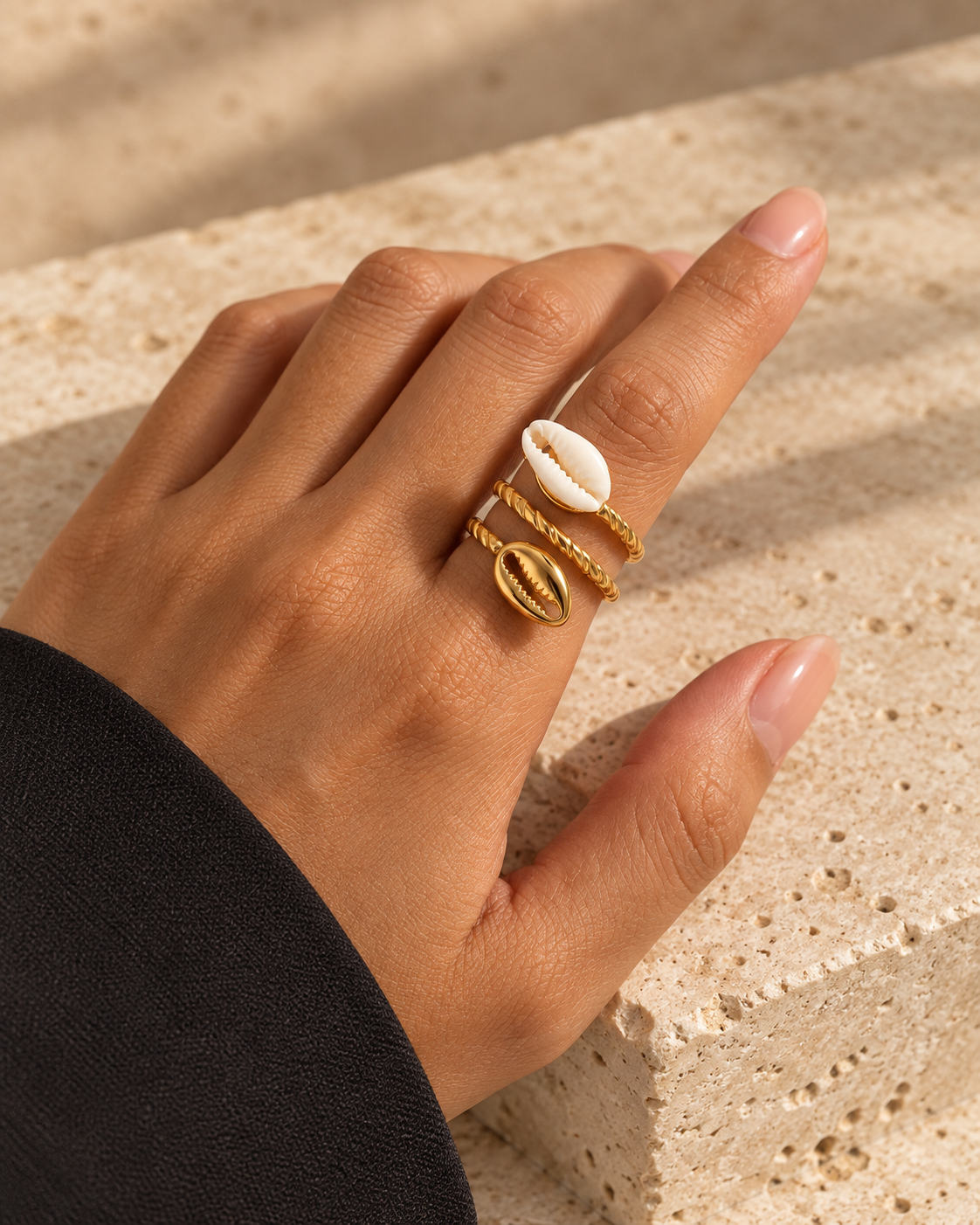 Bague COURA dorée à deux cauris, blanc et doré, portée à l’index sur fond de travertin beige.