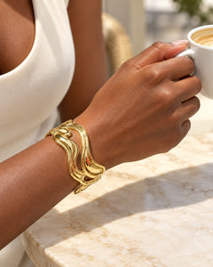 Bracelet Vague doré porté au poignet, avec une tenue crème, autour d’un café.